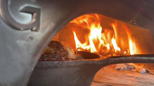 Chicken in a cast iron pan in the entrance of a Gozney Dome pizza oven with a wood fire behind it